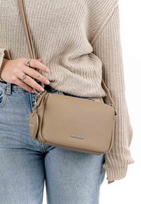 Beige leather crossbody bag with textured surface, silver hardware, and a heart-shaped accent tag, worn casually.