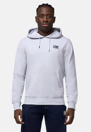Homme portant un sweat à capuche gris clair avec le logo "LEONE 1947" et un pantalon foncé, debout devant un fond uni.