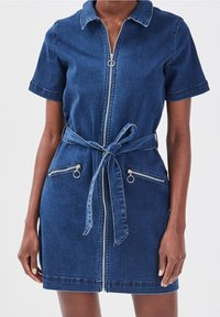 Robe en denim avec fermeture éclair sur le devant, manches courtes, taille cintrée par une ceinture, et deux poches zippées avec des accents en métal argenté. Couleur bleu foncé.