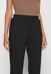 Detailní záběr osoby v černých kalhotách se šňůrkou a krajkovými bočními panely a bezrukávém hnědém žebrovaném topu.