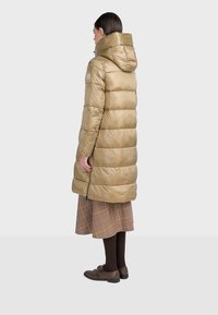 Lång vadderad jacka i ljusbrun färg med huva, quiltat mönster och sidofickor med dragkedja. Materialet har en blank, slät yta.