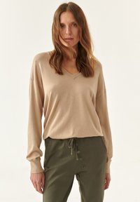 Suéter beige de cuello en V con mangas largas, combinado con pantalones verde oliva con cordón y detalles de botones en el costado. Textura suave.