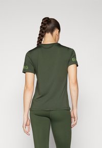 Oliwkowy t-shirt sportowy z krótkim rękawem, gładka tekstura, haftowane logo na lewym rękawie. Noszony z dopasowanymi legginsami.