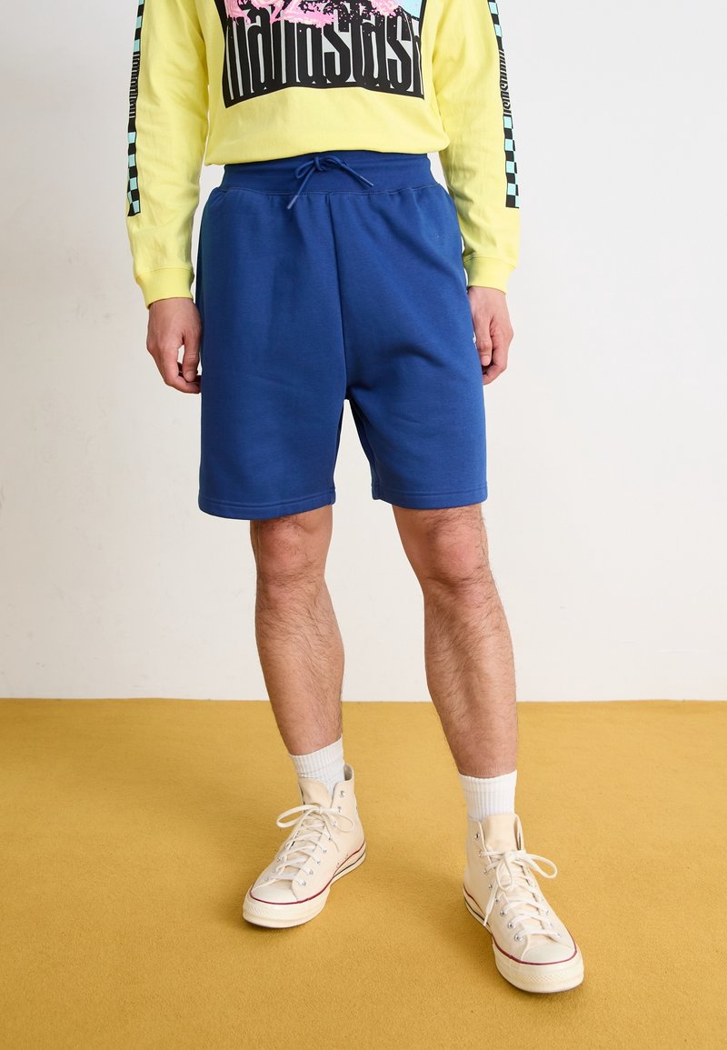 Personne portant un short bleu à cordon, des baskets montantes blanches et un t-shirt jaune à manches longues avec un motif noir et rose.