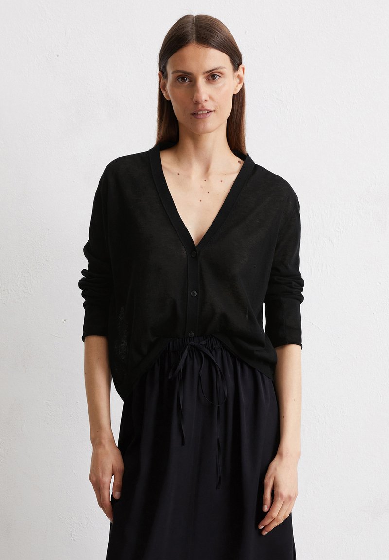 Schwarze Strickjacke aus leichtem Material, mit V-Ausschnitt, Knopfverschluss und Dreiviertelärmeln. Kombiniert mit einem schwarzen Rock mit Kordelzug.