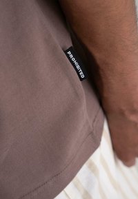 Brown cotton t-shirt with a visible black label reading "PROHIBITED" on the left side. Features a smooth texture and relaxed fit.