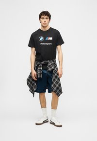 Νέος άνδρας που φορά μαύρο T-shirt BMW Motorsport, καρό πουκάμισο δεμένο γύρω από τη μέση, σορτς από τζιν, λευκές κάλτσες και λευκά αθλητικά παπούτσια.