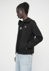 Μαύρο ελαφρύ μπουφάν με φερμουάρ μπροστά, κουκούλα και πλαϊνές τσέπες. Διαθέτει μικρό λογότυπο στο στήθος, φορεμένο πάνω από μαύρο μπλουζάκι.