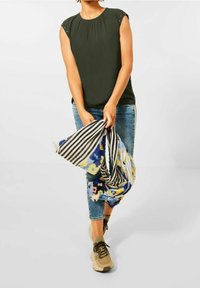 Tmavě zelený top s krátkým rukávem a plisovaným výstřihem, zdobený dekoračními prvky, kombinovaný s modrými džíny a šátkem s více vzory.