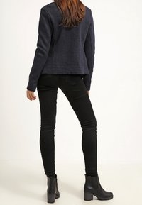 Veste polaire bleu marine à finition texturée, jean skinny noir et bottines à talons noires, vus de dos sur un fond uni.