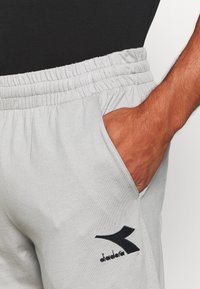 Närbild av en person som bär ljusgrå Diadora-träningsbyxor med elastisk midja, handen i sidofickan och en svart skjorta instoppad.