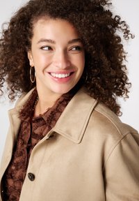 Jonge vrouw met krullend haar die glimlacht, draagt gouden oorringen, een bruine blouse met textuur en een beige jas met grote knopen.