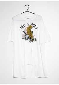 T-shirt bianco oversize con una grafica di una tigre gialla e fiori con il testo "FEEL STRONG" in nero. Maniche corte e tessuto leggero.