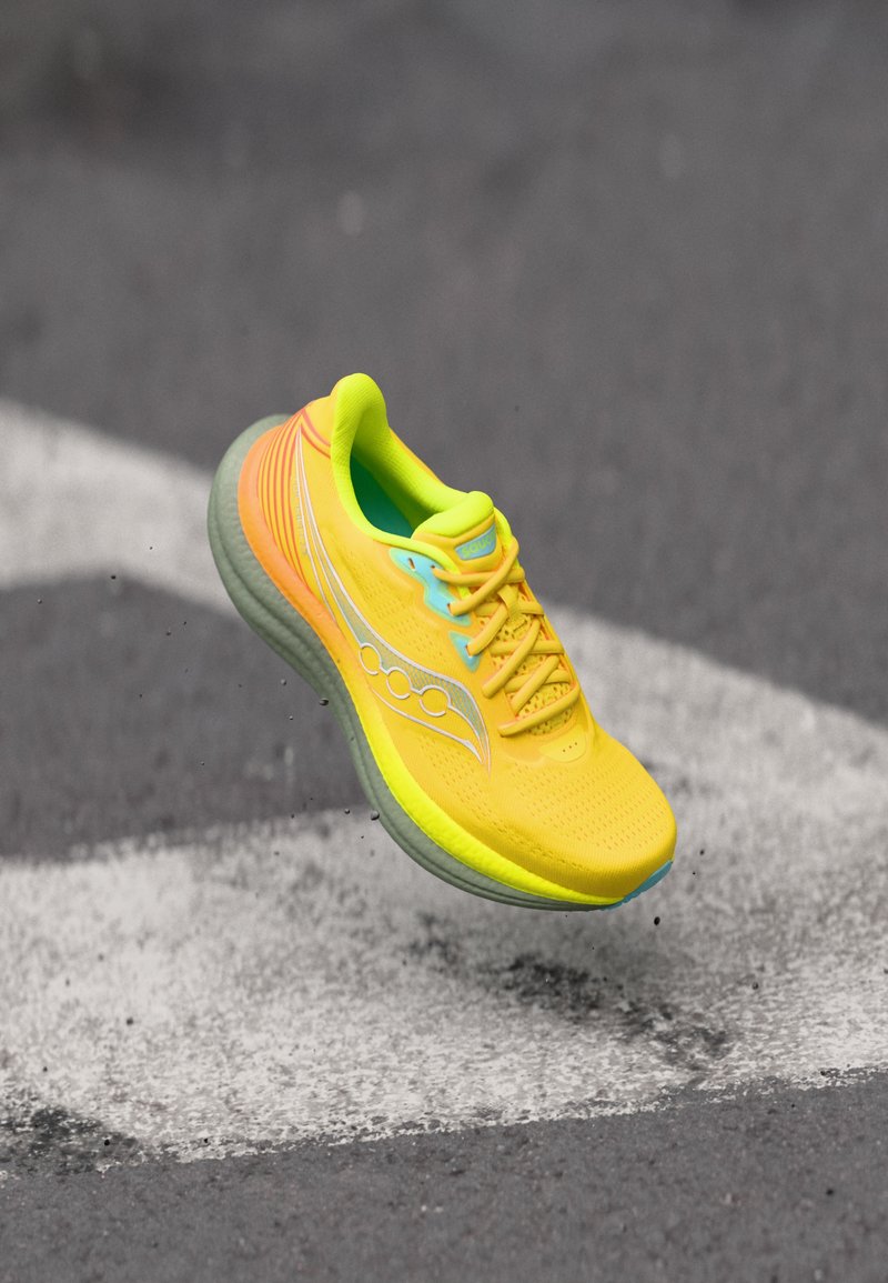 Φωτεινό κίτρινο παπούτσι για τρέξιμο με γκρι σόλα στον αέρα πάνω από άσφαλτο με λευκή βέλος.