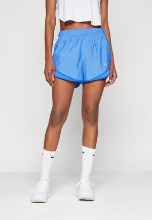 Kobieta ubrana w niebieskie sportowe spodenki Nike, białe skarpetki Nike i białe buty sportowe, stojąca na tle jednolitego jasnego tła.