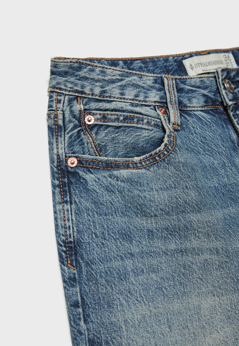 Jeans in denim blu che mostrano la tasca anteriore con rivetti di rame, passanti per cintura e una piccola etichetta del marchio in tessuto all'interno della vita.