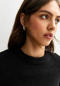Maglione nero lavorato a maglia con scollatura rotonda. Modella che indossa orecchini a cerchio in argento, caratterizzati da una finitura liscia e un design circolare.