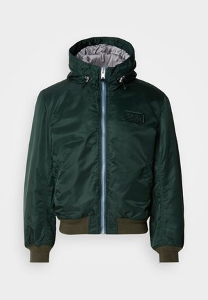 Giacca bomber verde scuro con cappuccio, foderatura interna argento, chiusura frontale con zip, polsini e orlo a costine, e patch con logo sul petto.