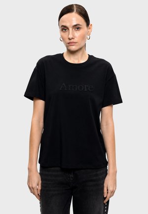 Donna con capelli raccolti indossa orecchini a cerchio dorati e una t-shirt nera con il testo "Amore" fatto di perline scure, in piedi davanti a uno sfondo chiaro e uniforme.