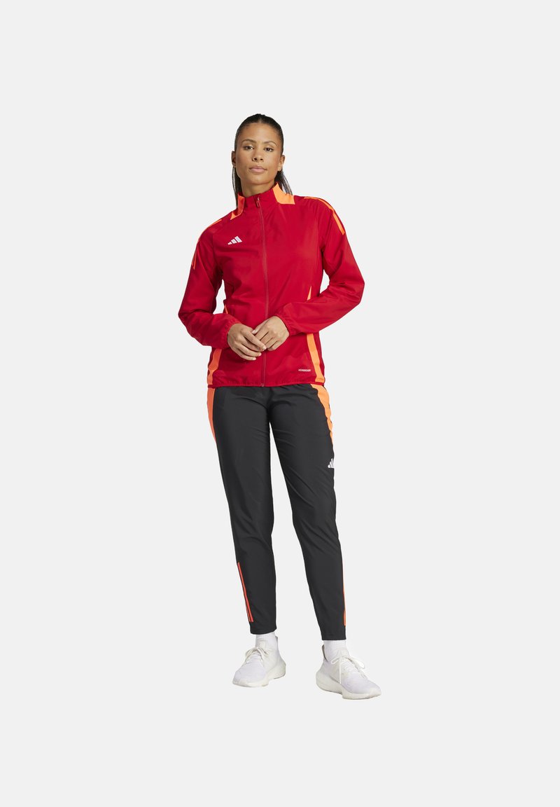 Veste zippée rouge avec des accents orange et pantalon noir avec des rayures orange, fabriqués en matériau léger. Logo Adidas sur les deux.