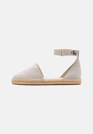 Espadrille de lona beige con tira para el tobillo, suela de yute y textura de tela suave. Presenta un logo en la tira. Diseño simple y minimalista.