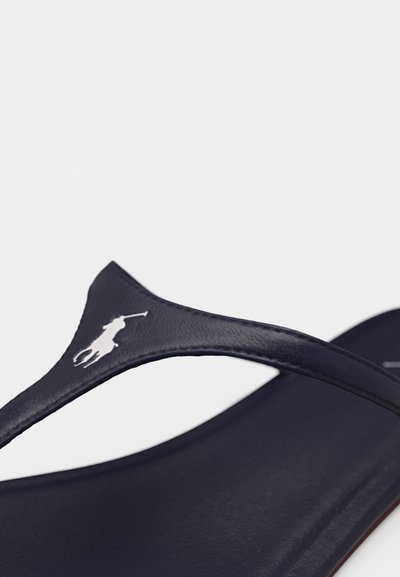 Κοντινό πλάνο από ιμάντα δερμάτινης σαγιονάρας σε σκούρο μπλε χρώμα με λογότυπο λευκού παίκτη πόλο σε απλό φόντο.