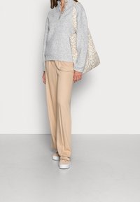 Grauer Rippstrickpullover mit hohem Kragen und drei Knöpfen, kombiniert mit beigen weit geschnittenen Hosen und einer gemusterten beigefarbenen Tragetasche. Weiße Sneakers.
