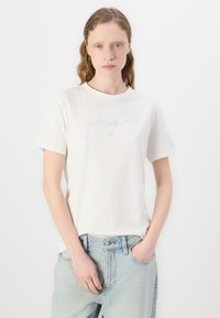 Tricou alb din bumbac cu mâneci scurte, având un logo subtil iridescent „Hugo” pe față, asortat cu blugi de culoare albastră deschisă.