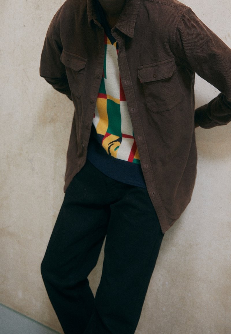 Braunes Cordhemd, zugeknöpft über einem mehrfarbigen, gemusterten Pullover. Schwarze Hose vervollständigt das Outfit. Hintergrund ist beige.