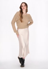 Beige gestrickter Pullover mit lockerer Passform und strukturiertem Detail. Cremefarbener Seiden-Midirock mit leichtem Saumverlauf. Schwarze Schnürstiefel.