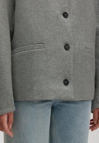 Grau knöpfige Jacke mit zwei Schlitztaschen, die über hellblauen Jeans getragen wird, während die Hände natürlich an den Seiten ruhen.