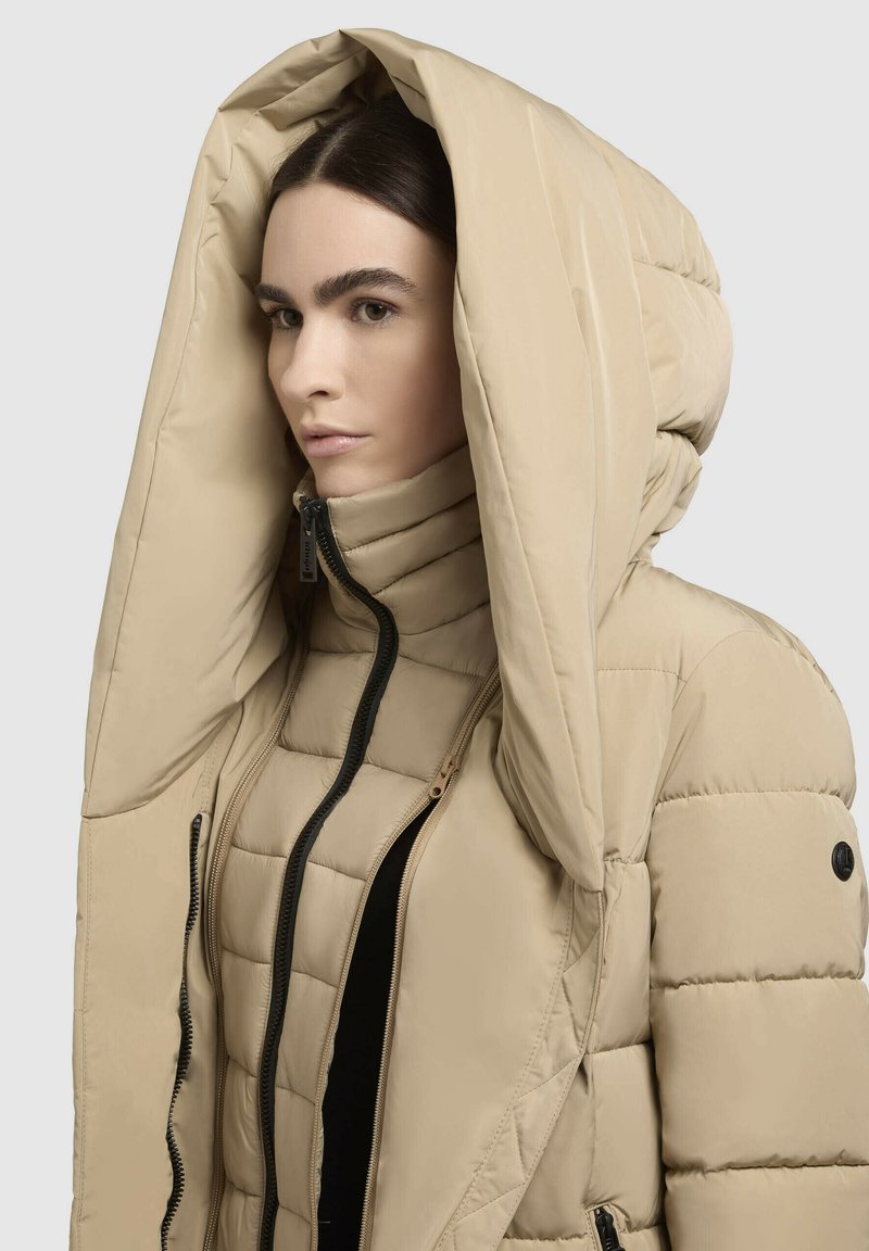 Frau in oversized beigefarbenem Pufferjacke mit großer Kapuze, hochgeschlossen am Hals, vor einfarbigem Hintergrund.