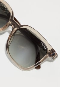 Ochelari de soare cu nuanță maronie, cu ramă transparentă, lentile gri și un accent metalic pe armătură. Prezintă logo-ul brandului "Vogue."