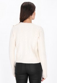 Crème kabelgebreide trui met een gestructureerd patroon, cropped lengte en een ronde halslijn. Beschikt over knoopaccenten op één schouder.