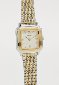 Montre carrée avec un bracelet en acier inoxydable argent et or, dotée d'un cadran blanc, d'accents dorés et d'un chiffre romain à la position 12 heures.