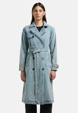 Kvinne iført lys blå denim trenchcoat med belte i midjen og svarte knapper, stående mot en ensfarget hvit bakgrunn.