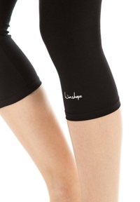 Melnas līdz ceļiem leggingas, kas izgatavotas no elastīga auduma, ar gludu tekstūru un nelielu balto logo "Winshape" tuvumā apakšmalai.