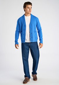 Blauer Cardigan aus weichem Strickstoff mit V-Ausschnitt, Knopfleiste und zwei Taschen. Kombiniert mit dunkelblauer Jeanshose und braunen Schuhen.