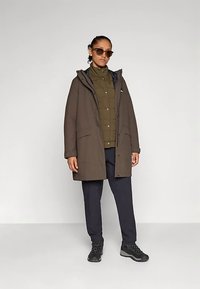 Brown parka jacket with a hood, featuring front pockets and a zip closure, paired with an olive green shirt and navy pants. Black hiking boots.