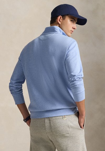 Férfi világoskék hosszú ujjú ingben, szürke nadrágban, sötétkék sapkában és órával, kezét a zsebében tartva, oldalra nézve áll.