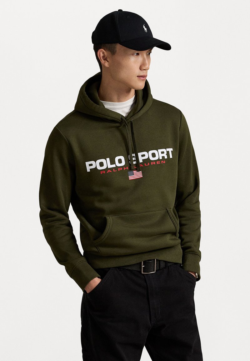 Olīvzaļa kapuce ar tekstu "POLO SPORT" un "RALPH LAUREN", priekšējā kabata, apvienota ar melniem biksēm un melnu cepuri ar logotipu.