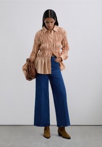 Kvinna i en rynkad persikofärgad blus, vidbeniga blåjeans, bruna ankelstövlar, håller en brun läderväska, står mot en grå bakgrund.