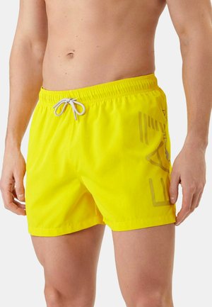 Mandlig torso iført lyse gule badeshorts med grå snor og subtilt sidegrafik, stående mod en ensfarvet lys baggrund.