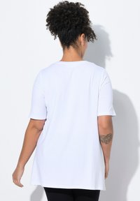 Camisa blanca de manga corta con cuello redondo, hecha de una tela suave. Presenta un dobladillo trasero más largo y un tatuaje floral en la parte superior del brazo.