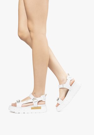 White platform sandals with an open toe design, featuring adjustable straps, metallic accents, and a textured sole. Brand name visible.