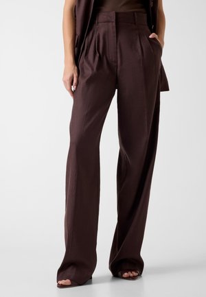 Kobieta nosząca ciemnobrązowe spodnie o wysokim stanie i szerokich nogawkach z plisami oraz buty na otwartym palcu, stojąca z jedną ręką w kieszeni.