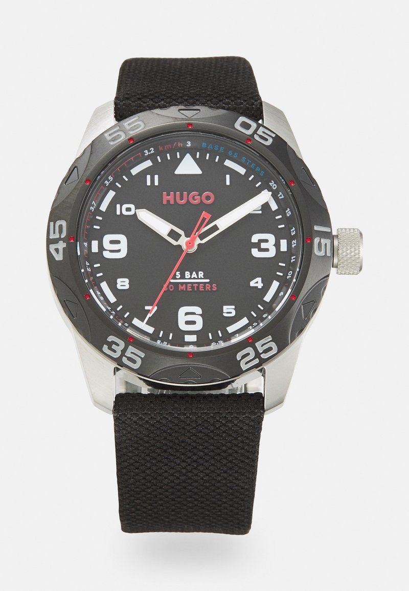 Reloj negro con caja redonda de acero inoxidable, esfera oscura, números blancos y acentos rojos. Presenta una correa de tejido negro texturizado.