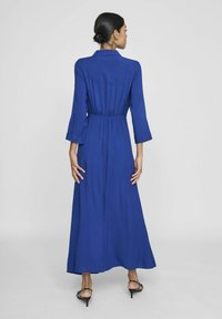 Ilgai mėlyna suknelė su apykakle, elastingu juosmens srityje ir ilgomis rankovėmis. Lygus tekstūrinis paviršius ir plūdenos dizainas. Suknelė turi pilną ilgį sijoną.