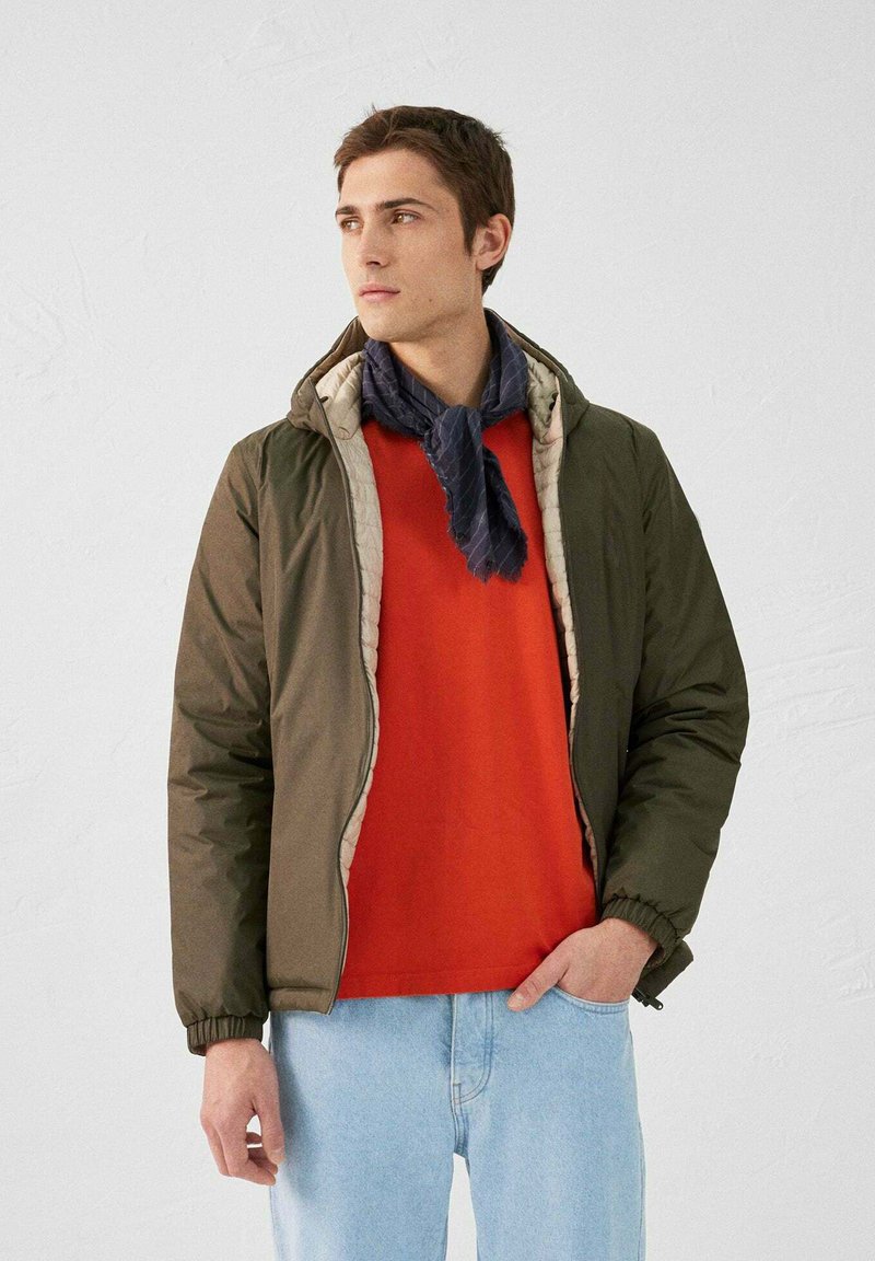 Doudoune vert olive avec une doublure beige, portée sur un sweat-shirt rouge. Une écharpe à motifs bleu foncé est nouée autour du cou. Jeans bleu clair.