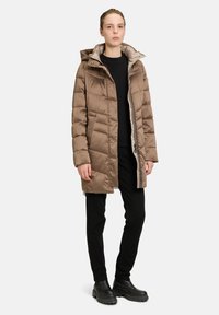 Braune isolierte Daunenjacke mit Kapuze, mit gesteppten Details, einem Reißverschluss vorne und seitlichen Taschen. Getragen über einem schwarzen Oberteil und Hosen.